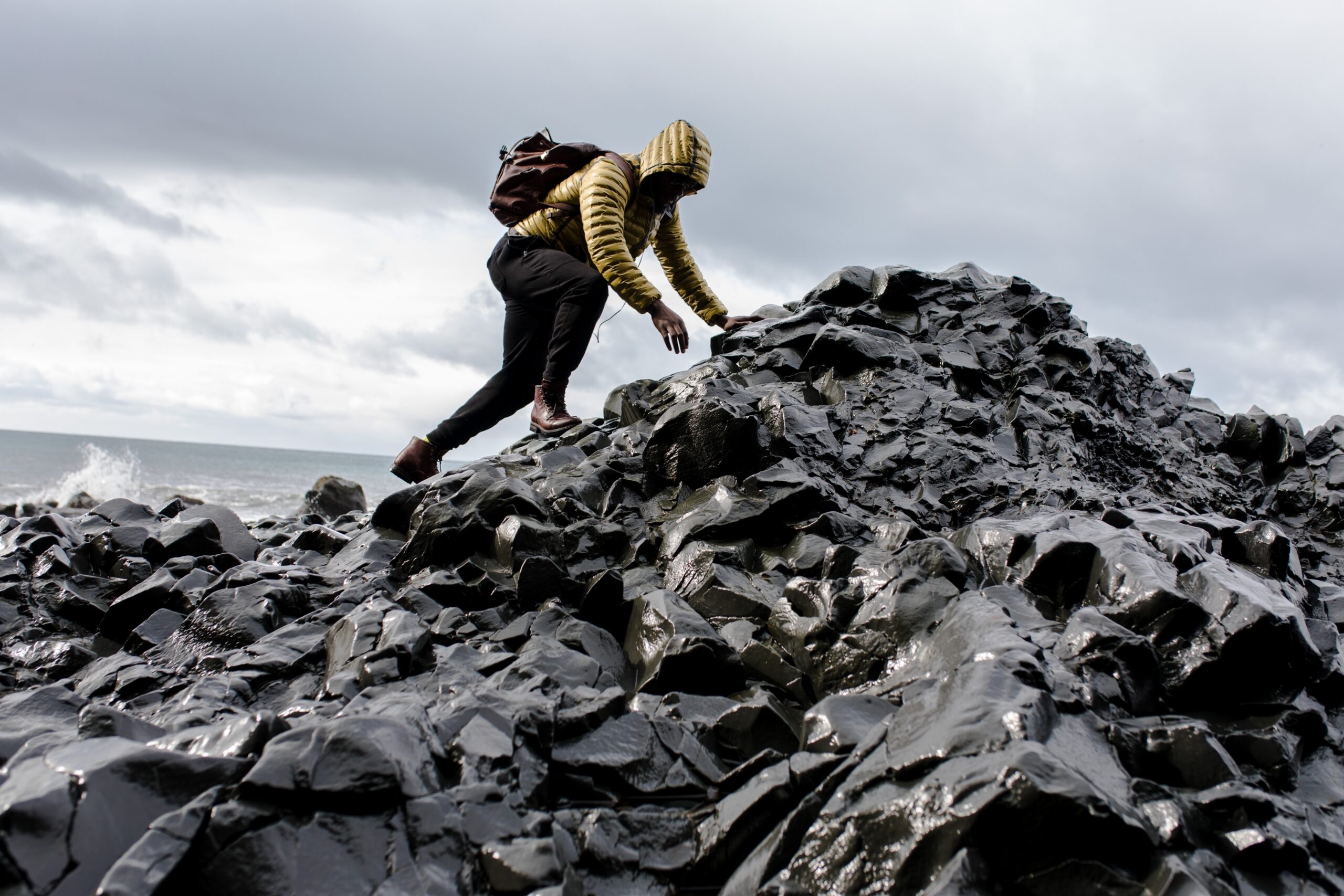 Man in yellow hiking jacket and black trousers scrambles up large pile of rocks